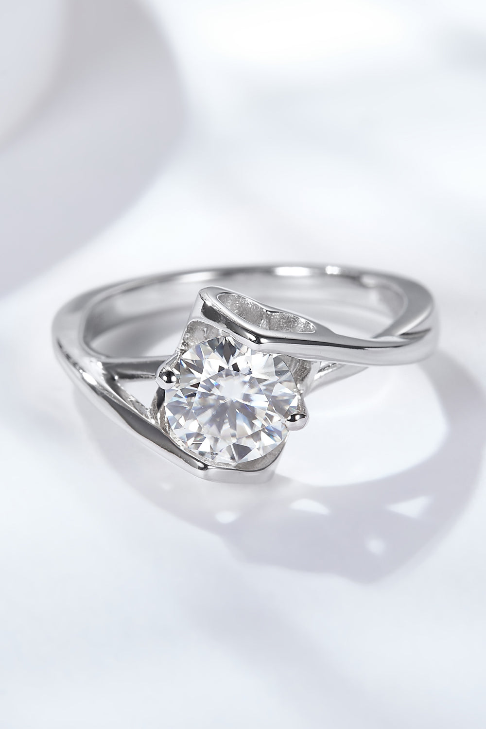 Top-down view of a 1 ct round moissanite solitaire ring on a white satin background, prong setting and slender band visible.