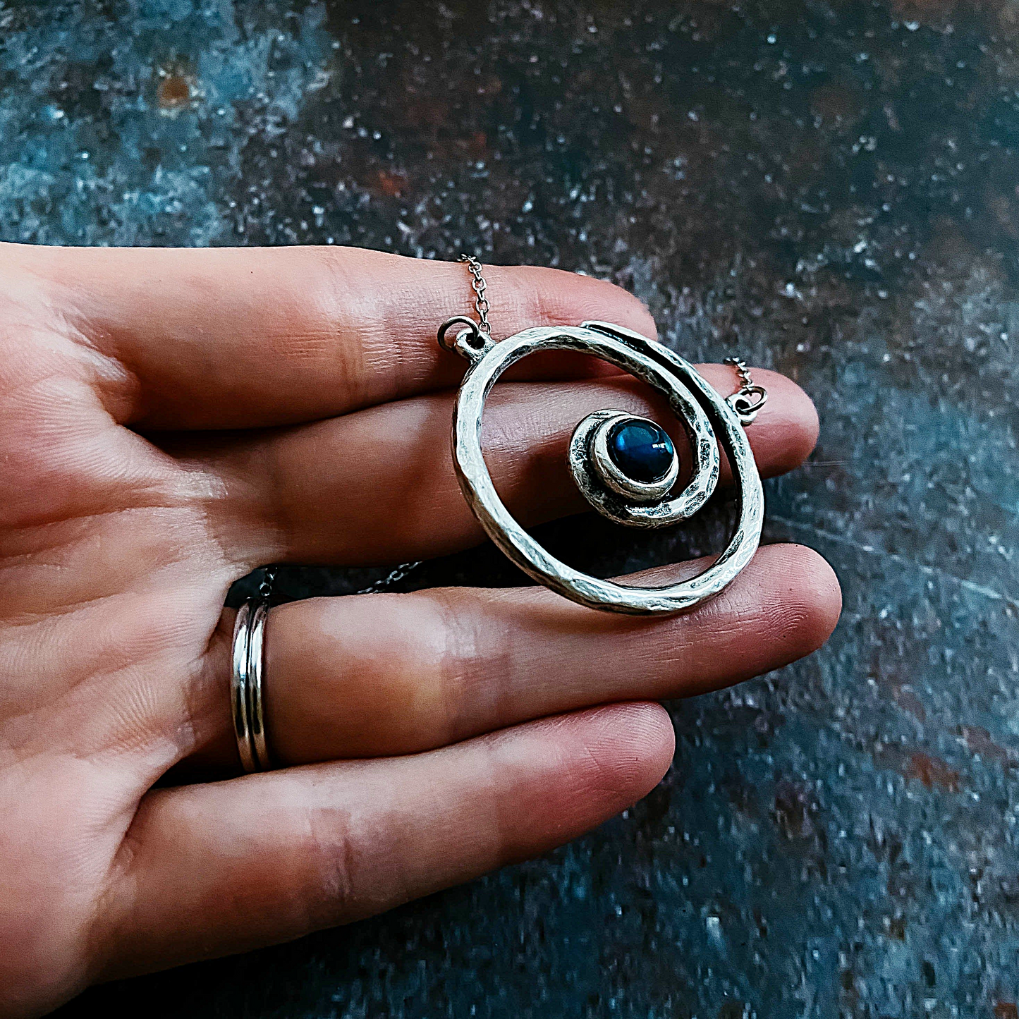 Hand holding a silver pendant with a blue stone against a textured dark background