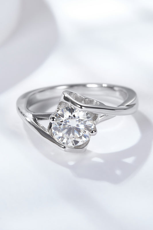 Top-down view of a 1 ct round moissanite solitaire ring on a white satin background, prong setting and slender band visible.