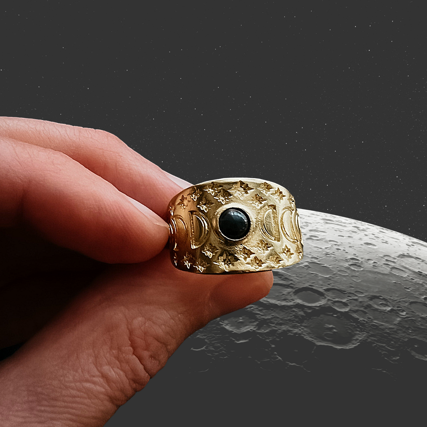 Gold ring with a black gemstone held by a hand against a moon-like surface