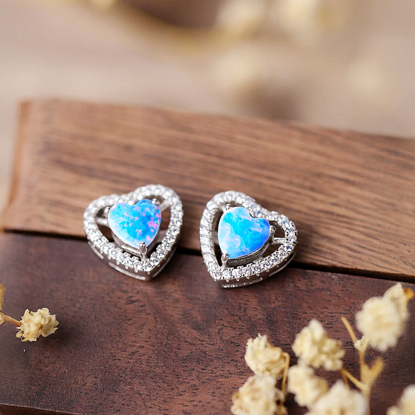 Heart-shaped earrings with blue gemstones on a wooden surface with dried flowers.